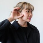 Portrait d'une femme asiatique avec la main sur l'oreille, symbolisant l'écoute ou l'attention, sur fond blanc.