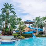 Piscine avec toboggan bleu entouré de palmiers et de parasols sous un ciel nuageux