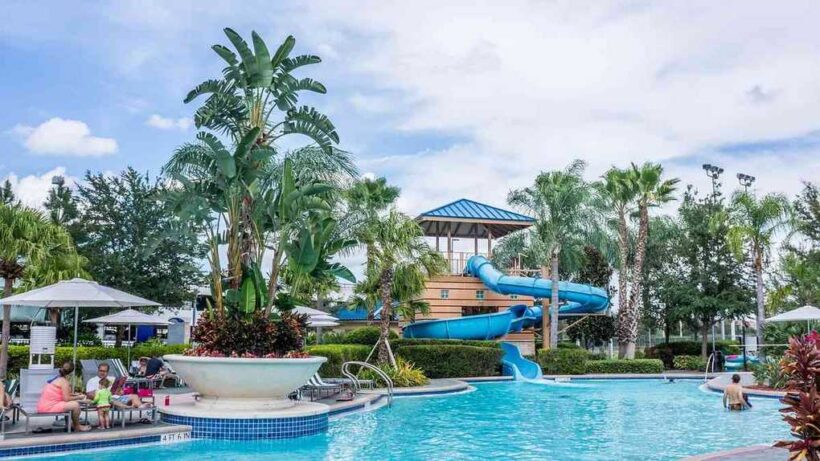 Piscine avec toboggan bleu entouré de palmiers et de parasols sous un ciel nuageux